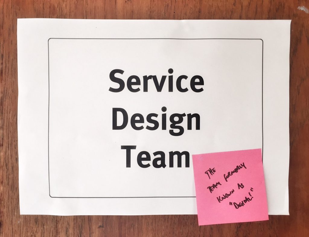 a sign saying 'service design team' with a stickly note on it saying 'the team formerly known as 'digital''