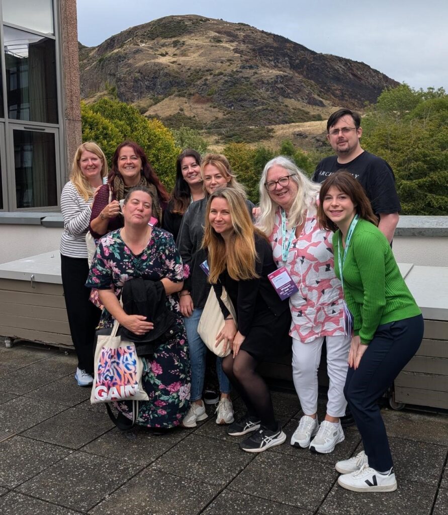 9 of the 10 blog authors posing and smiling at SDinGov in Edinburgh, with Arthur's Seat (a famous hill) in the background