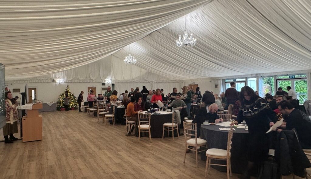 Alt text: A large room with chandeliers and a draped white ceiling hosting multiple round tables where people are seated and standing, engaging in conversation. A decorated Christmas tree is visible in the background, and a speaker stands near a podium at the front.