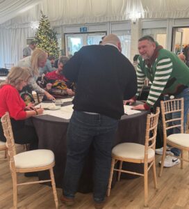 A group of people gathered around a round table, writing on large sheets of paper during a collaborative activity. Chairs surround the table, and a decorated Christmas tree is visible in the background.