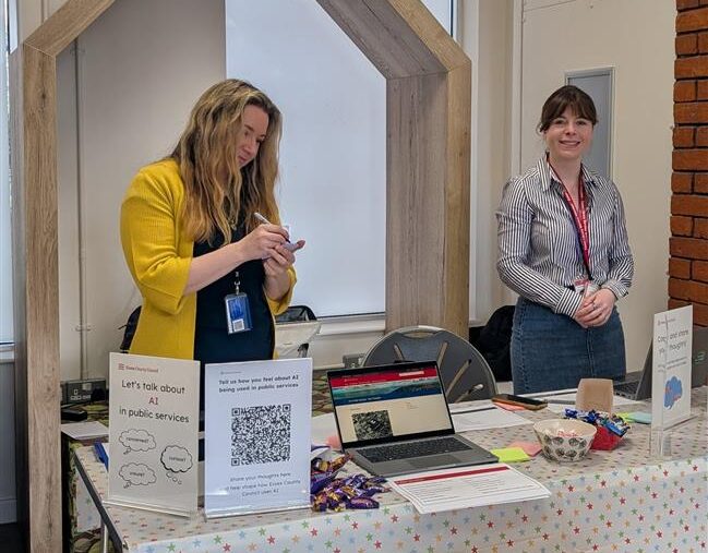 Two white women in a library standing behind a desk, with signs inviting people to take part in research activities.